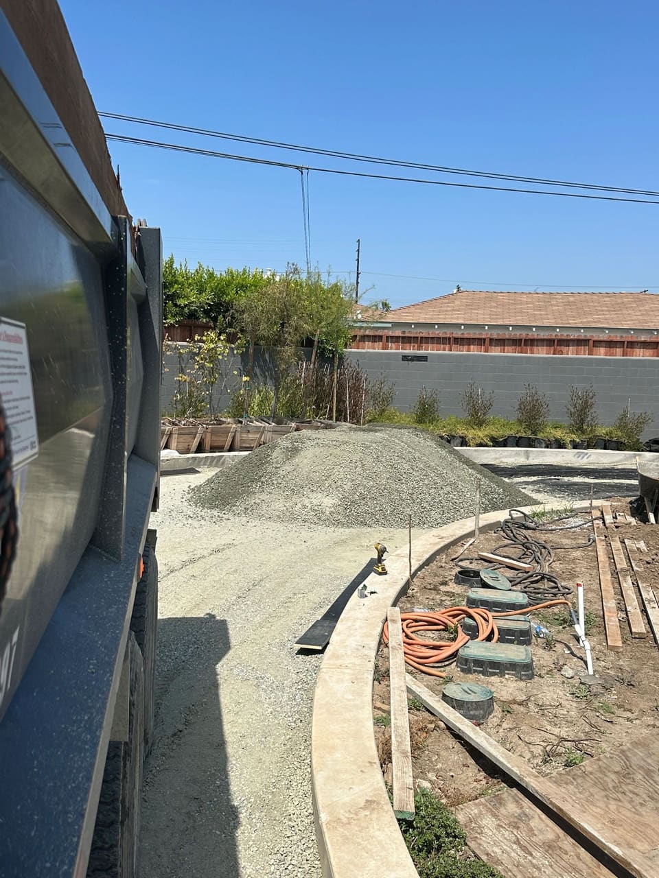 A pile of gray gravel sits in a backyard under construction beside a curved concrete curb and a dump truck.