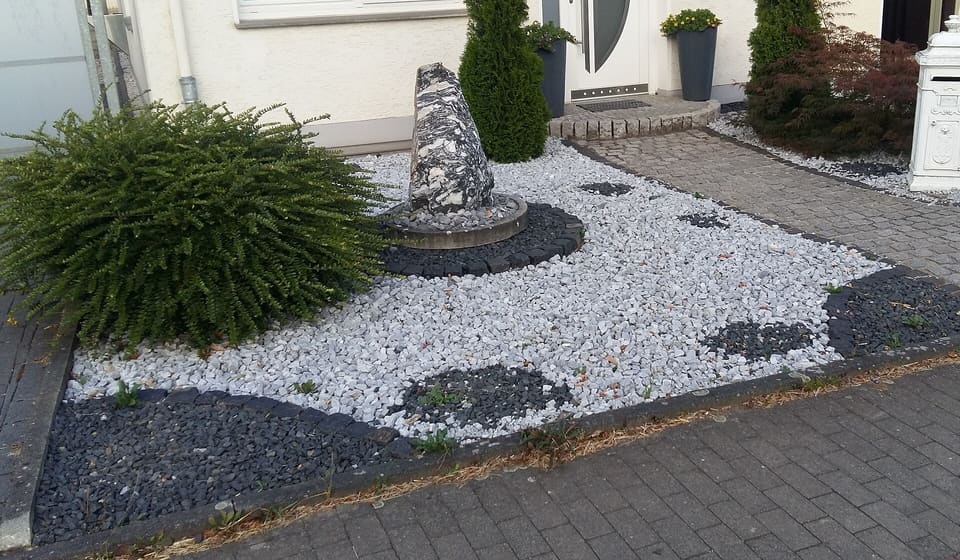 A front-yard bed of white marble chippings with islands of dark gray basalt gravel around a standing stone and a clipped shrub, beside a paved driveway.