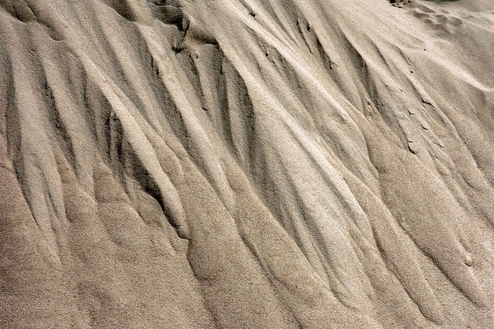 Close-up of the sloping face of a pile of fine, light tan sand with shallow flow grooves running diagonally down the surface.